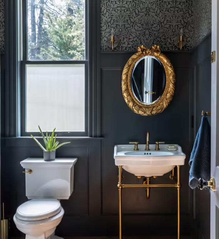 Powder room with dark floral wallpaper, paneled wainscot, a brass console sink, and an oval gilt mirror