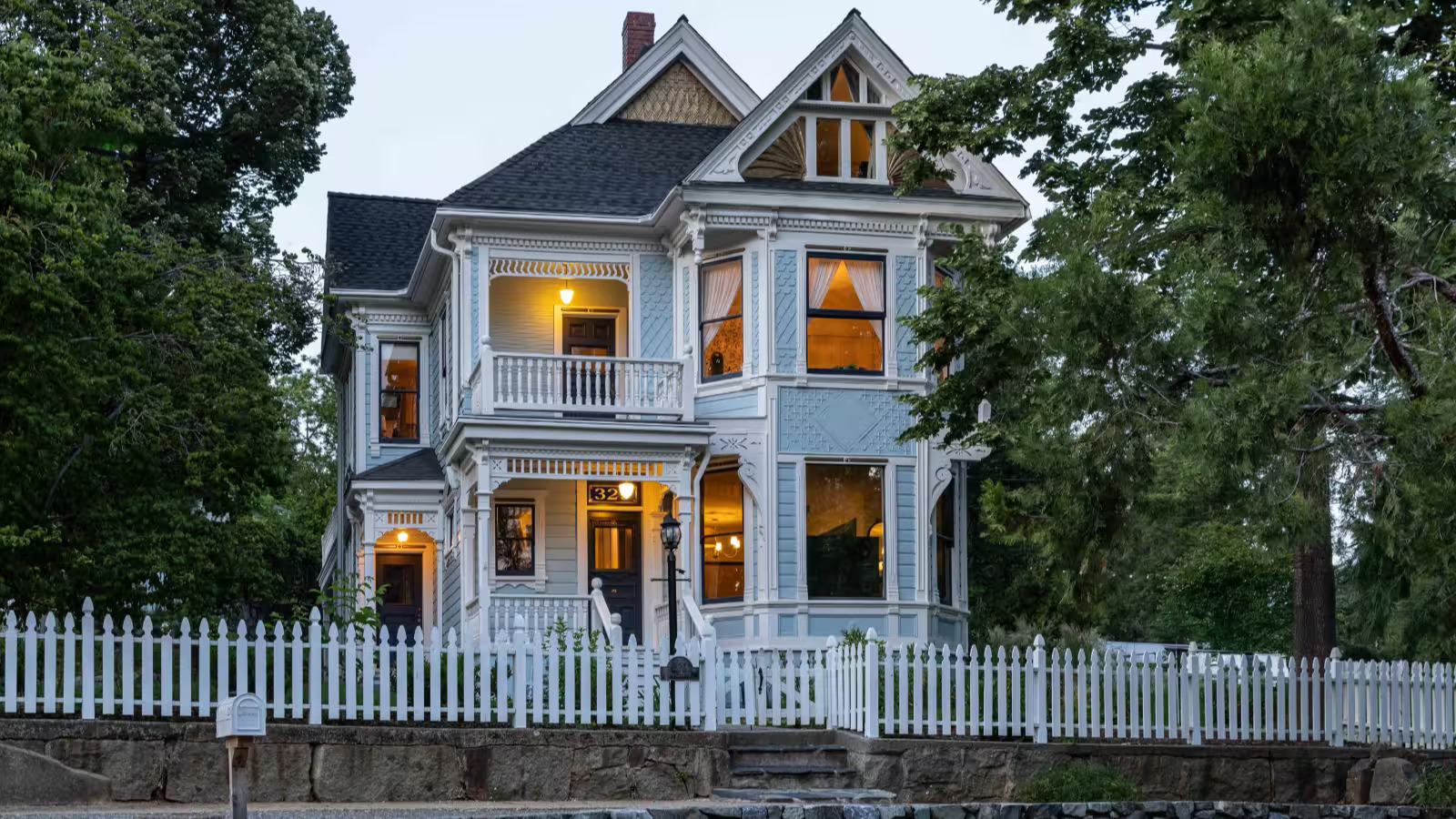 Restored three-story Victorian lit at dusk behind a picket fence and stone retaining wall in Nevada City